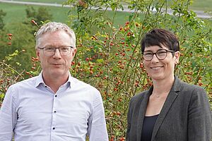 Ein Mann und eine Frau posieren draussen vor einem grünen Hintergrund.