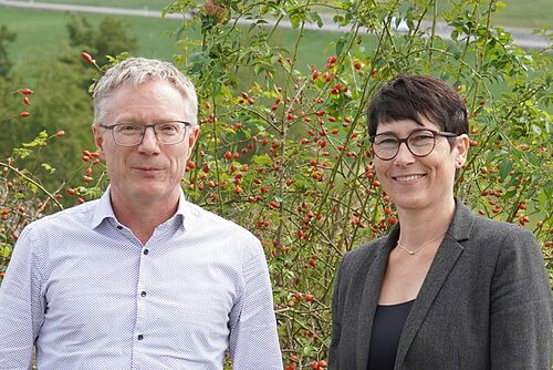 Ein Mann und eine Frau posieren draussen vor einem grünen Hintergrund.