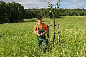 Ein Mann in Arbeitskleidung steht neben einem jungen Baum auf einer Wiese. 