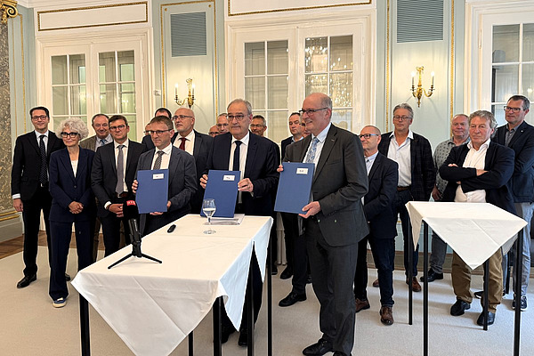 Eine Gruppe von Männern und einer Frau in einem Saal mit gedeckten Tischen.
