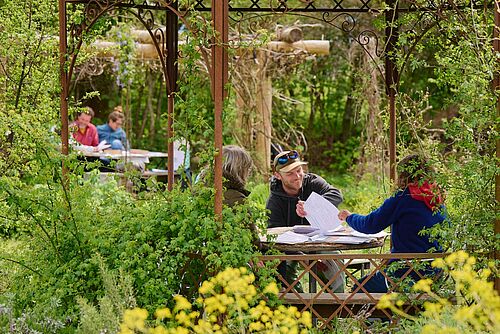 Eine Gruppe Menschen sitzen an einem Tisch unter einem Pavillon in einem üppig wuchernden, grünen Garten. Sie halten Unterlagen in den Händen und besprechen sich.