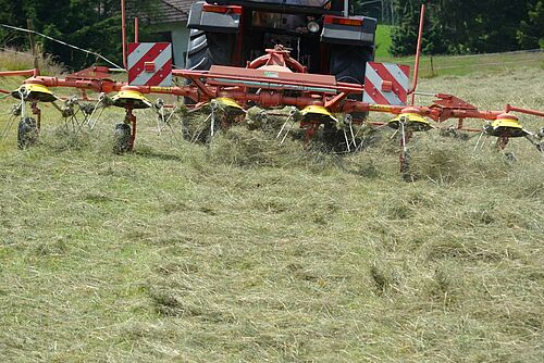 Auf dem Bild ist ein Heuschüttler an einem Traktor zu sehen, im Vordergrund liegt getrocknetes Heu auf einer Wiese. 