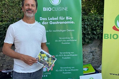 Mann steht lächelnd vor einer grünen Fahne und hält das Abzeichen von Bio Cuisine in der Hand