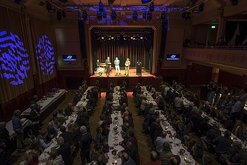 Ein gut besetzter Saal von oben, hinten eine Bühne mit mehreren Personen drauf.