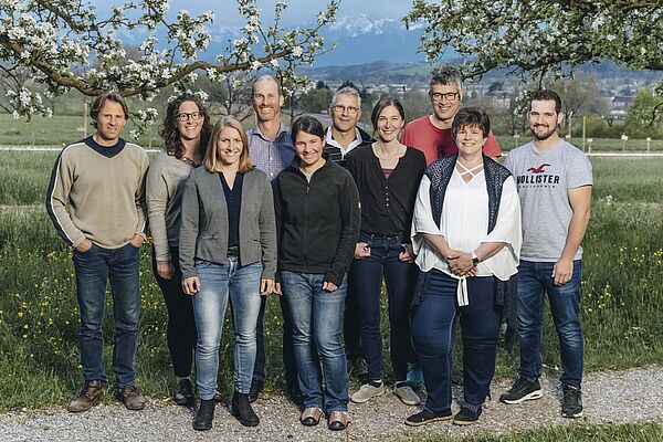 Eine Menschengruppen posiert vor einem Feld unter zwei blühenden Bäumen. 