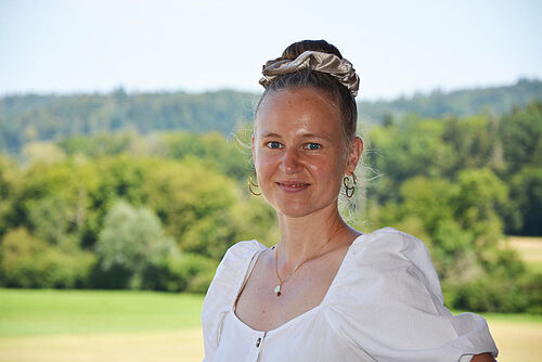 Eine Frau mit hochgesteckten Haaren vor bewaldetem Hintergrund.