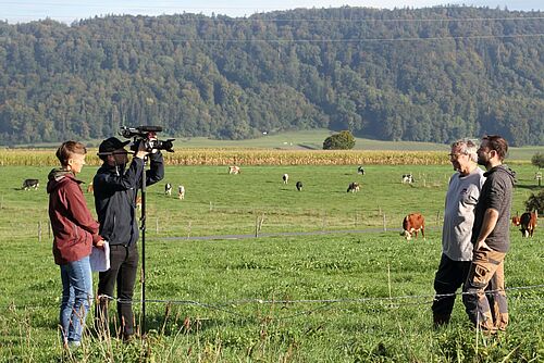 Ein Kamaeramann und eine Kollegin interviewen in einem Feld zwei Männer. 