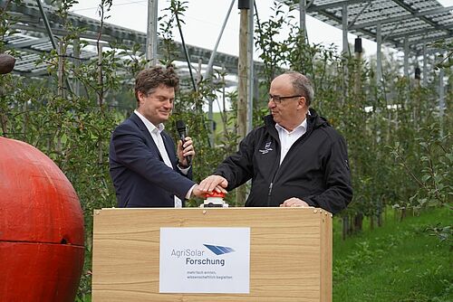 Zwei Männer stehen auf einem Podium und drücken einen grossen roten Knopf, um die Solaranlage zu starten. 